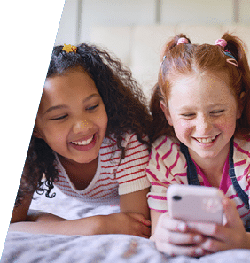 Three cheerful young girls lie on a textured bedspread, engrossed in a smartphone. Their diverse hairstyles are adorned with colorful clips and ties, radiating joy and camaraderie.
