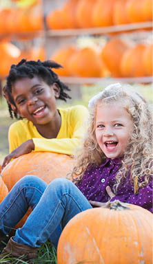 A group of three multi-ethnic children, 4-5 years old, sitting on the ground with lots of pumpkins at a market stand, smiling at the camera.